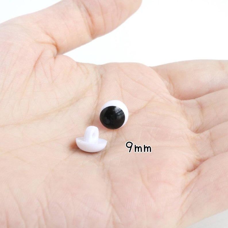 Op de afbeelding: Twee kleine, ronde witte plastic knutselogen met zwarte pupillen, elk met een diameter van 9mm. E&eacute;n oog is geplaatst om de gladde, bolle voorkant te tonen, terwijl de andere is gedraaid om de witte oogbevestiging aan de achterkant te laten zien.