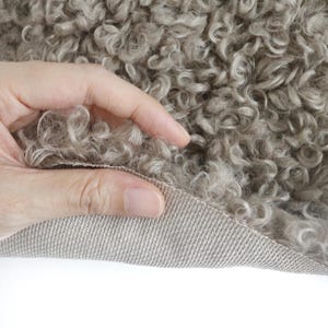 May include: A piece of taupe-colored curly faux fur fabric with a dense, textured pile. The material features a woven, light-colored backing visible where the fabric is folded, showcasing the contrast between the soft, curly surface and the base.