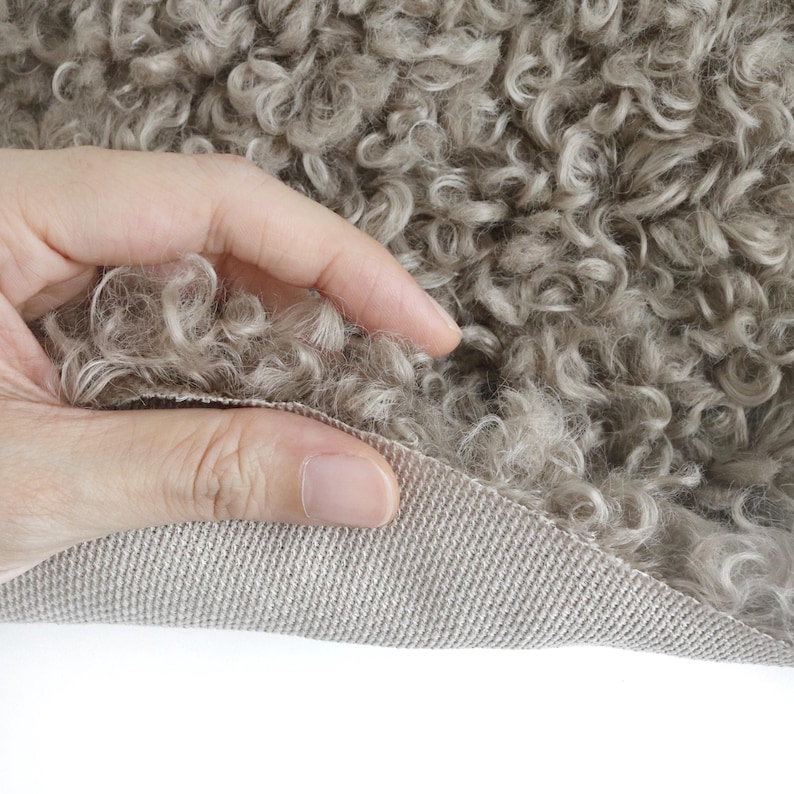 May include: A piece of taupe-colored curly faux fur fabric with a dense, textured pile. The material features a woven, light-colored backing visible where the fabric is folded, showcasing the contrast between the soft, curly surface and the base.