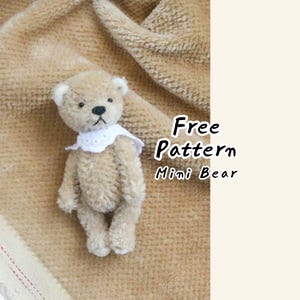 May include: A small beige teddy bear with dark eyes and a black nose, wearing a white eyelet lace collar. The text Free Pattern Mini Bear is displayed to the right of the bear. The bear has a fuzzy texture and is positioned on a tan fabric.