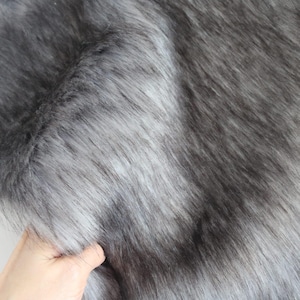 Puede incluir: Primer plano de una tela de piel sintética gris. La tela tiene fibras largas y suaves en diferentes tonos de gris, creando una apariencia texturizada. El material se está sosteniendo, mostrando su flexibilidad y caída. Ideal para manualidades o decoración del hogar.