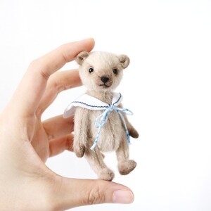 K&ouml;nnte beinhalten: Ein beigefarbener Miniatur-Teddyb&auml;r mit dunklen Augen und einer schwarz gestickten Nase. Der B&auml;r tr&auml;gt einen wei&szlig;en Matrosenkragen mit dunkelblauem Saum und ein hellblau-wei&szlig; kariertes Band, das zu einer Schleife um den Hals gebunden ist.