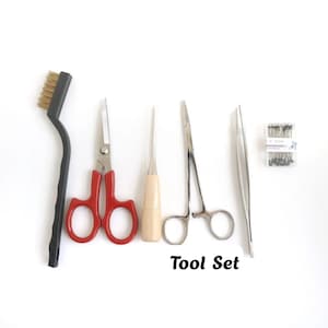 May include: A tool set consisting of a black wire brush, scissors with red handles, an awl with a light wooden handle, silver metal locking forceps, silver tweezers, and a small clear plastic container filled with black pins labeled Tool Set.