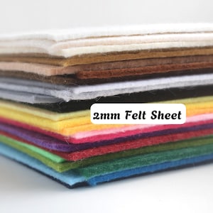 Peut inclure: Une pile de feuilles de feutrine rectangulaires de différentes couleurs, notamment blanc, beige, marron, gris, noir, jaune, rose, violet, vert et bleu. Le texte 2mm Felt Sheet est imprimé sur une étiquette blanche sur le côté de la pile.