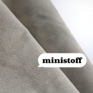 May include: Close-up of two rolls of light gray fabric with a textured surface. The fabric is tightly rolled, revealing the texture and color variations. The word "ministoff" is in a white speech bubble.