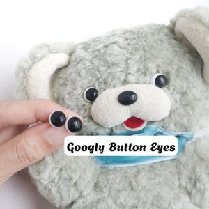 Op de afbeelding: Een knuffel met lichtgroene krullende vacht, een witte snuit, een rode tong en een lichtblauw lint om de nek. Het speelgoed heeft zwart-witte wiebelogen. De tekst "Googly Button Eyes" is in het zwart op een witte achtergrond gedrukt.
