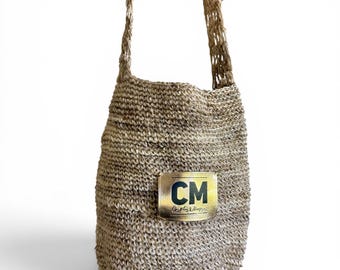 May include: A tan, woven straw shoulder bag with a textured, open-knit appearance. The bag features a rectangular gold-toned metallic patch on the front, displaying the bold black letters CM and the smaller text Chapitas & Mapas.