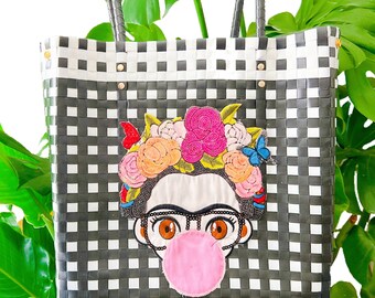 May include: Black and white checkered tote bag with a Frida Kahlo-inspired design. The bag features a beaded floral crown, butterfly accents, and a bubblegum bubble. The tote has black handles and gold rivets.