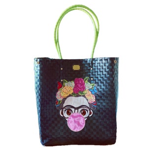 May include: Black woven tote bag with a Frida Kahlo-inspired design. The bag features a colorful floral crown, butterfly accents, and a pink bubble gum bubble. It has light green handles and gold-colored hardware.