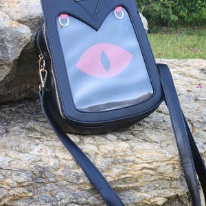Puede incluir: Bolso bandolera negro con diseño de orejas de gato. El bolso presenta un panel frontal transparente con un gráfico de ojo de gato rosa. Tiene cierre de cremallera, herrajes dorados y una correa larga y ajustable. El bolso está sobre una roca.