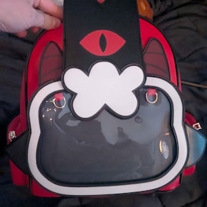 Può includere: Zaino rosso e nero con un design a forma di gatto. Presenta una parte superiore nera con un occhio rosso, un accento bianco a forma di nuvola e una tasca frontale trasparente. Il testo "ACTUAL BAG" è visibile.