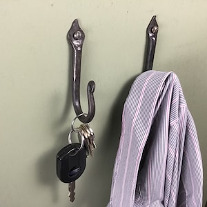 May include: Two black metal hooks mounted on a green wall. One hook has a set of keys and a car key attached to it. The other hook has a striped shirt hanging from it.