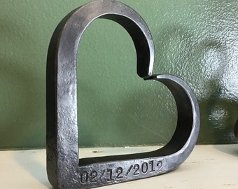 May include: A dark gray, heart-shaped metal sculpture. The sculpture has a rough, textured surface and the date "02/12/2012" is stamped on the bottom. The heart is displayed on a white surface against a green background.