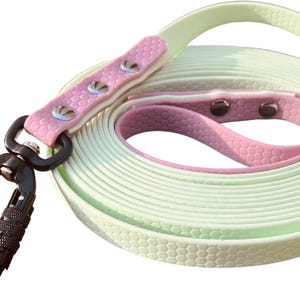 Könnte beinhalten: Eine lange, einziehbare Hundeleine mit einem schwarzen Karabinerhaken. Die Leine besteht aus zwei Farben Gummi, einem hellgrünen und einem rosa. Die Leine hat eine strukturierte Oberfläche.
