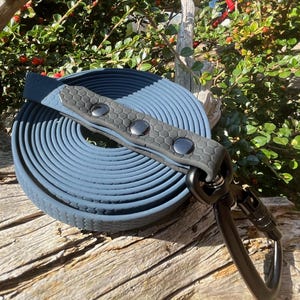 Op de afbeelding: Een opgerolde blauwe hondenriem met een grijze handgreep met een honingraatpatroon en drie zilveren klinknagels. De riem heeft een zwarte metalen clip. De achtergrond omvat groen en een houten oppervlak.
