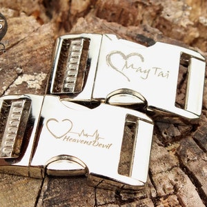 May include: Two silver metal dog collar buckles with engraved hearts and text. The top buckle has the text "May Tai" and the bottom buckle has the text "Heavens Devil" with a heartbeat symbol.