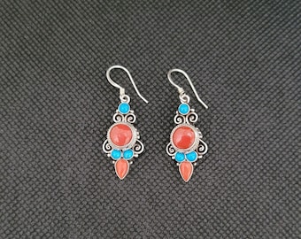 May include: A pair of silver dangle earrings with red and blue gemstone accents. The earrings feature a round red stone surrounded by smaller blue stones.