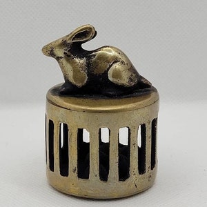 May include: A small, antique brass container with a rabbit figurine on top. The container has a cylindrical shape with vertical cutouts. The rabbit is in a resting position, adding a touch of whimsy to the piece.