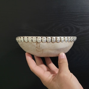 May include: A white ceramic bowl with a silver rim decorated with a row of small, round, silver skull-shaped accents.