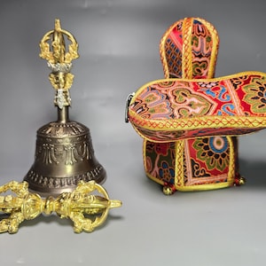 May include: A bronze-colored metal bell with a gold-toned handle, a gold-toned vajra scepter, and a rectangular fabric cushion featuring a vibrant, multi-colored floral and geometric pattern with gold trim and a zipper closure.