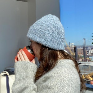 May include: A person wearing a light blue knitted beanie and a light gray fuzzy sweater is holding a red travel mug and taking a sip. The person is sitting at a table with a white canvas tote bag.
