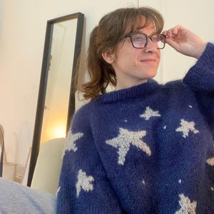 May include: A navy blue jumper featuring white star designs. The jumper has long sleeves and a round neck. The person in the image is wearing glasses and jeans.