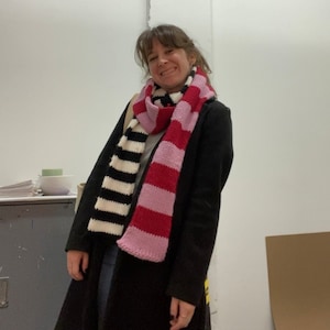 May include: A woman wearing a black coat with a striped scarf. The scarf is black and white on one side and pink and red on the other side.