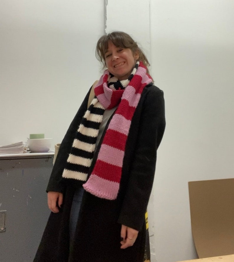 May include: A woman wearing a black coat with a striped scarf. The scarf is black and white on one side and pink and red on the other side.