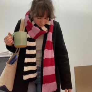 May include: A woman wearing a black coat and a striped scarf with pink and red stripes. She is holding a green ceramic mug.