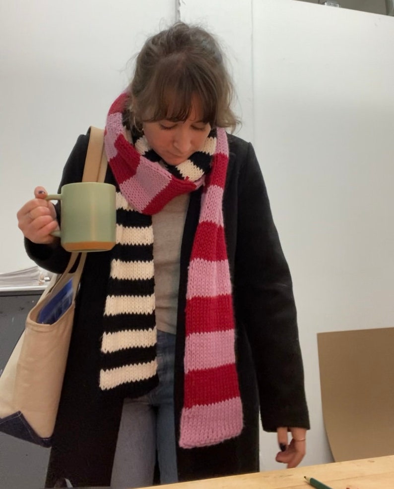 May include: A woman wearing a black coat and a striped scarf with pink and red stripes. She is holding a green ceramic mug.