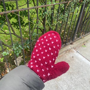 Puede incluir: Un mitón rojo tejido con un patrón de pequeños corazones blancos. El mitón está hecho de un hilo suave y texturizado. El mitón se muestra sobre un fondo de una valla y vegetación.