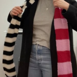 May include: A person wearing a black coat with two striped scarves, one black and white and the other pink and red.