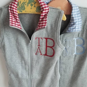 May include: Two gray sleeveless vests with contrasting colored collars. The vest on the left has a red and white gingham collar and the vest on the right has a blue and white gingham collar. Both vests have embroidered initials on the front. The vest on the left has the initials "T.A.B." embroidered in red and the vest on the right has the initials "T.M.B." embroidered in blue.