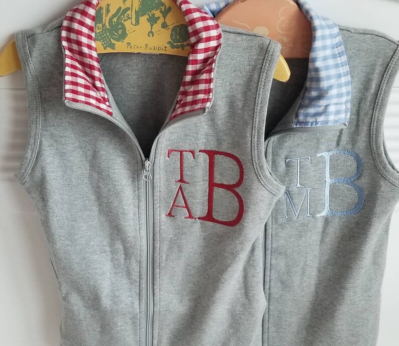 May include: Two gray sleeveless vests with contrasting colored collars. The vest on the left has a red and white gingham collar and the vest on the right has a blue and white gingham collar. Both vests have embroidered initials on the front. The vest on the left has the initials "T.A.B." embroidered in red and the vest on the right has the initials "T.M.B." embroidered in blue.