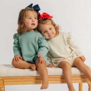 May include: Two long-sleeved knit rompers with ruffled shoulder details. One romper is sage green and the other is cream. The sage green romper is paired with a dark navy blue hair bow, and the cream romper is paired with a bright red hair bow.