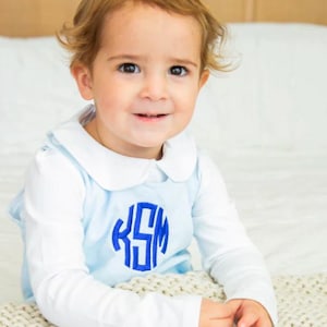 May include: A baby wearing a light blue and white long-sleeved collared shirt with a blue monogram that says "KSM" on the front. The baby is sitting on a white knitted blanket.