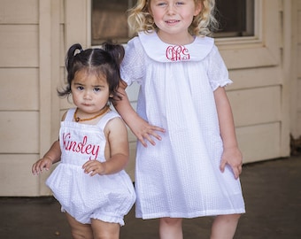 May include: Two young girls wearing white smocked dresses with red embroidered monograms. The girl on the left is wearing a white smocked romper with the name "Kinsley" embroidered in red. Both girls are wearing red shoes.