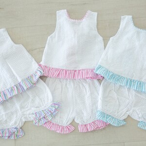 Puede incluir: Tres blusas blancas sin mangas con dobladillos con volantes y bombachos a juego. Las blusas tienen volantes de diferentes colores: rosa, azul y multicolor. Los bombachos tienen volantes de colores a juego.