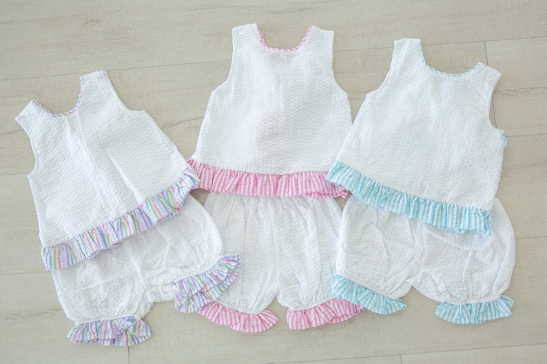 Puede incluir: Tres blusas blancas sin mangas con dobladillos con volantes y bombachos a juego. Las blusas tienen volantes de diferentes colores: rosa, azul y multicolor. Los bombachos tienen volantes de colores a juego.