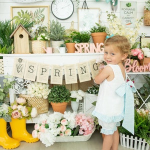 Puede incluir: Una ni&ntilde;a peque&ntilde;a con un vestido blanco y un lazo azul y blanco a rayas se encuentra frente a un estante blanco con una pancarta de primavera y varias plantas y flores. El estante tiene las palabras "Bloom" y "Spring".