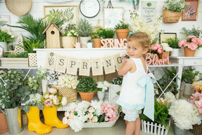 Puede incluir: Una ni&ntilde;a peque&ntilde;a con un vestido blanco y un lazo azul y blanco a rayas se encuentra frente a un estante blanco con una pancarta de primavera y varias plantas y flores. El estante tiene las palabras "Bloom" y "Spring".