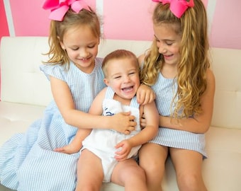May include: Two girls wearing blue and white striped dresses with pink bows in their hair are sitting on a white couch with a younger boy wearing a white onesie. The girls are smiling and the boy is laughing.