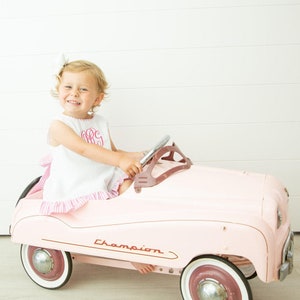 Puede incluir: Una ni&ntilde;a peque&ntilde;a con un vestido rosa y blanco con un monograma rosa conduce un coche de juguete rosa con un volante cromado. El coche tiene la palabra "Champion" impresa en el lateral.