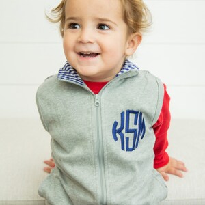 May include: A grey zip-up vest with a blue monogram "KSM" on the front. The vest has a blue and white checkered collar and is worn over a red long-sleeved shirt.