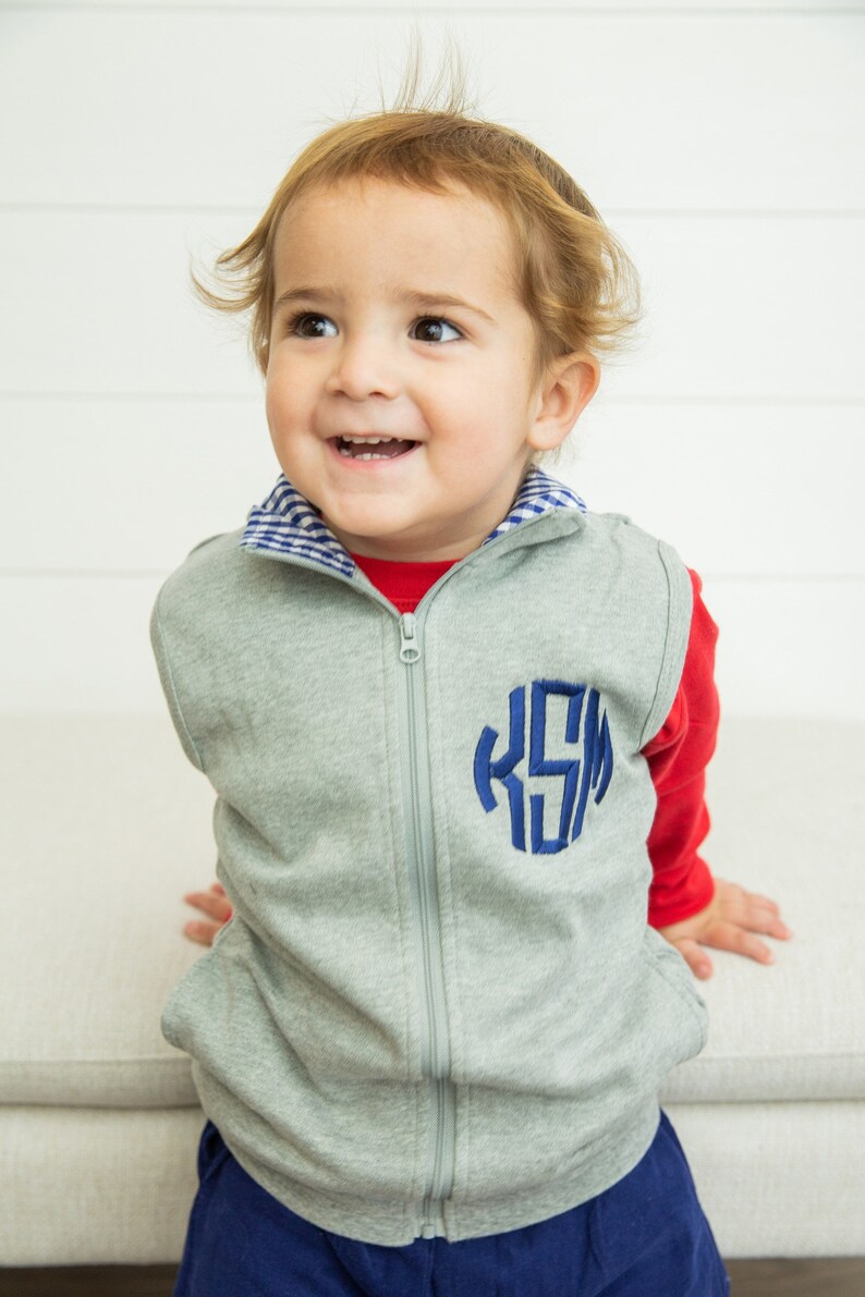 May include: A grey zip-up vest with a blue monogram "KSM" on the front. The vest has a blue and white checkered collar and is worn over a red long-sleeved shirt.