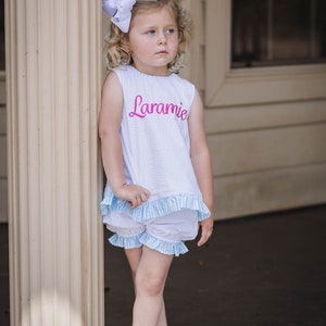 Puede incluir: Una ni&ntilde;a peque&ntilde;a con un top blanco sin mangas con ribete de vichy azul y blanco y pantalones cortos a juego. El top tiene el nombre "Laramie" bordado en rosa en la parte delantera. Lleva un lazo blanco en el pelo y sandalias blancas.