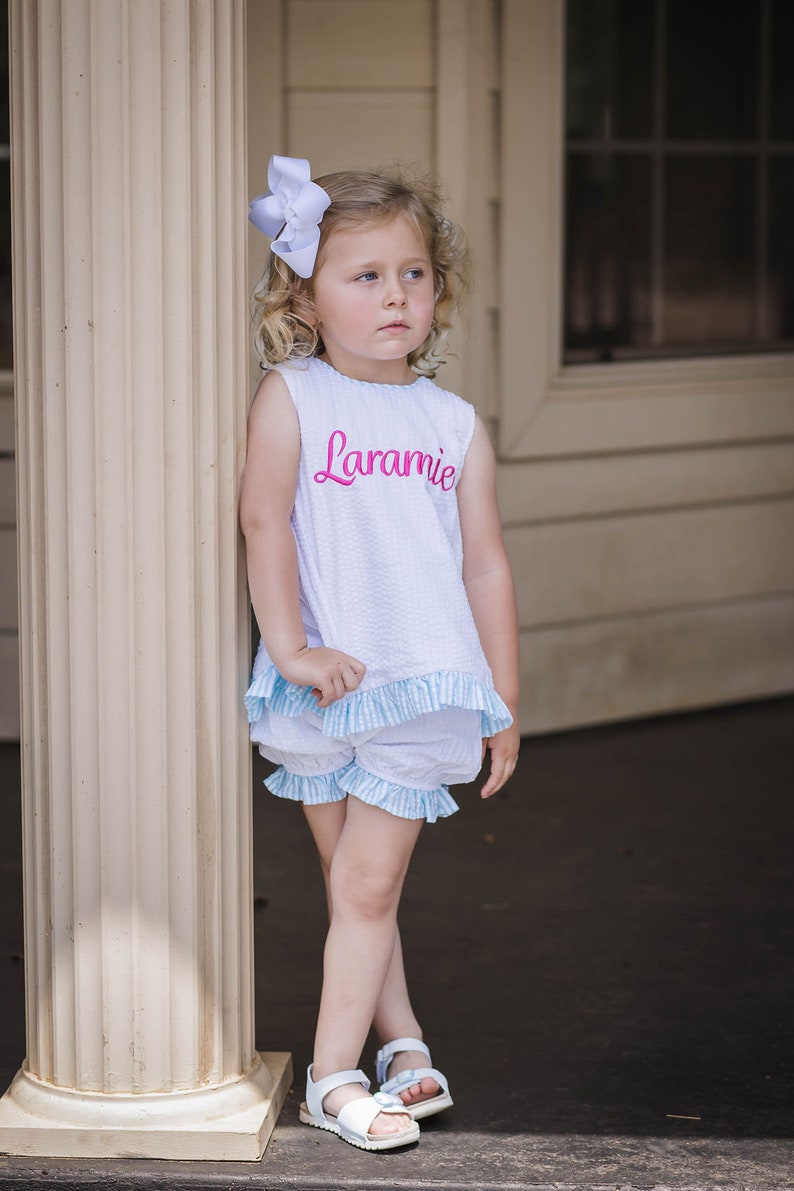 Puede incluir: Una ni&ntilde;a peque&ntilde;a con un top blanco sin mangas con ribete de vichy azul y blanco y pantalones cortos a juego. El top tiene el nombre "Laramie" bordado en rosa en la parte delantera. Lleva un lazo blanco en el pelo y sandalias blancas.