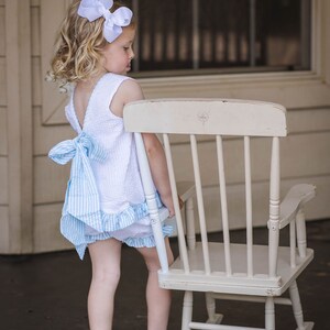 Puede incluir: Una ni&ntilde;a peque&ntilde;a con un vestido blanco y azul de cuadros vichy y un lazo blanco en el pelo est&aacute; de pie junto a una mecedora blanca.