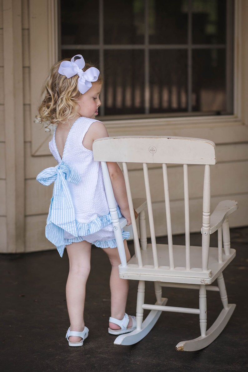 Puede incluir: Una ni&ntilde;a peque&ntilde;a con un vestido blanco y azul de cuadros vichy y un lazo blanco en el pelo est&aacute; de pie junto a una mecedora blanca.