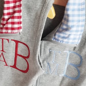 May include: Two gray sweatshirts with a zipper closure. One sweatshirt has a red and white gingham collar and the embroidered letters "T A B". The other sweatshirt has a blue and white gingham collar and the embroidered letters "T M B".
